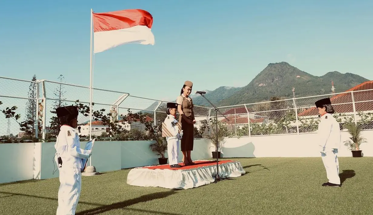 Bertindak sebagai pemimpin upacara, Yuni Shara berdiri di panggung berukuran mini dengan karpet merah yang dikelilingi kain berkerut putih polos. Ia menghadap mikrofon. Di belakang, ada bendera merah putih berkibar. Para petugas upacara adalah murid PAUD. Mereka berbaju putih plus kupluk hitam. Upacara yang digelar di rooftop ini terasa khidmat. (Foto: Dok. Instagram @yunishara36)