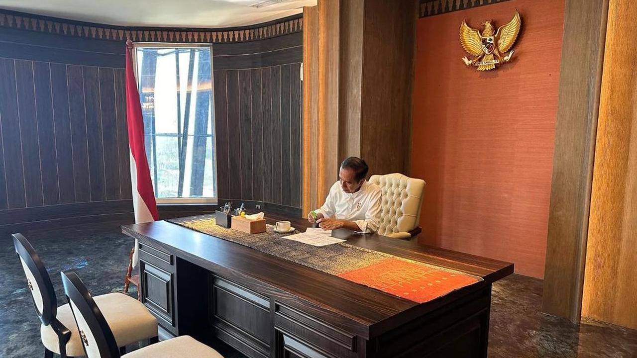 Pada foto pertama, Jokowi tampak duduk di belakang meja yang cukup besar di ruang kerja. Dia terliat memeriksa sejumlah dokumen di atas mejanya sambil memegang pulpen. Secangkir minuman tampak menemaninya.