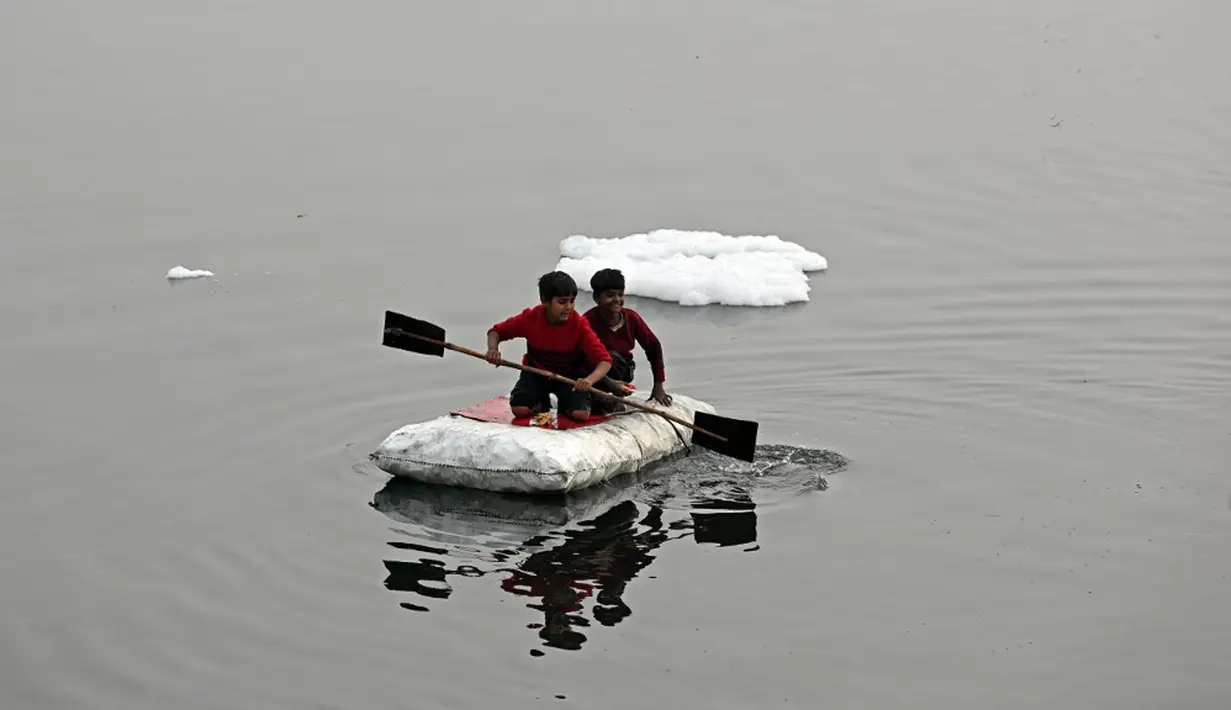 Anak-anak mendayung rakitnya sambil mengumpulkan bahan-bahan daur ulang dekat potongan busa tercemar yang mengambang di Sungai Yamuna, New Delhi, India, 26 Desember 2021. Busa yang menyengat itu mengandung amonia dan fosfat tingkat tinggi. (Sajjad HUSSAIN/AFP)