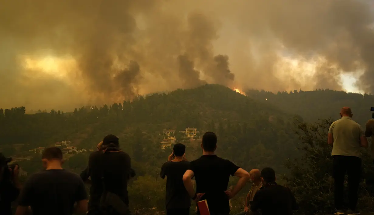 Penduduk setempat berdiri di atas bukit saat api mendekat di desa Gouves di pulau Evia, sekitar 185 kilometer (115 mil) utara Athena, Yunani, Minggu (8/8/2021). Pilar asap dan abu yang mengepul mengubah langit berwarna jingga di atas pulau terbesar kedua di Yunani. (AP Photo/Petros Karadjias)