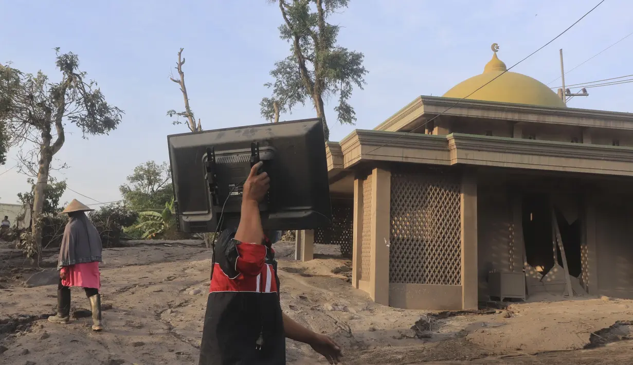 Seorang pria membawa televisi saat berjalan melewati masjid yang sebagian terkubur abu vulkanik dari letusan Gunung Semeru di desa Kajar Kuning di Lumajang, Jawa Timur, Indonesia, Senin, 5 Desember 2022. Gunung Semeru mengalami kenaikan dari Level III (Siaga) menjadi Level IV (Awas) sejak 4 Desember 2022 pada pukul 12.00 WIB, sehingga Pusat Vulkanolologi dan Mitigasi Bencana Geologi memberikan beberapa rekomendasi agar masyarakat mematuhinya. (AP Photo/Imanuel Yoga)