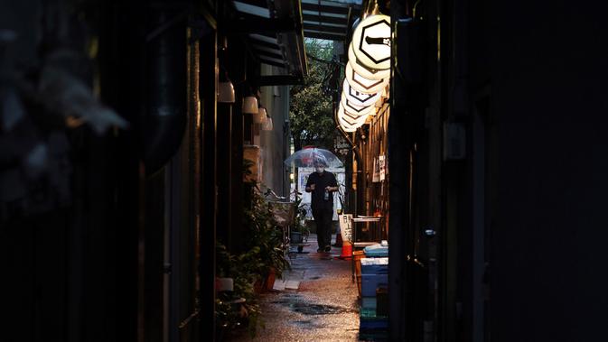 Seorang pria yang mengenakan masker untuk membantu mencegah penyebaran COVID-19 berjalan melewati gang sempit saat hujan di Tokyo, Jepang, Selasa (30/6/2020). Sebagian besar kegiatan sosial dan bisnis di Jepang telah kembali dimulai. (AP Photo/Eugene Hoshiko)