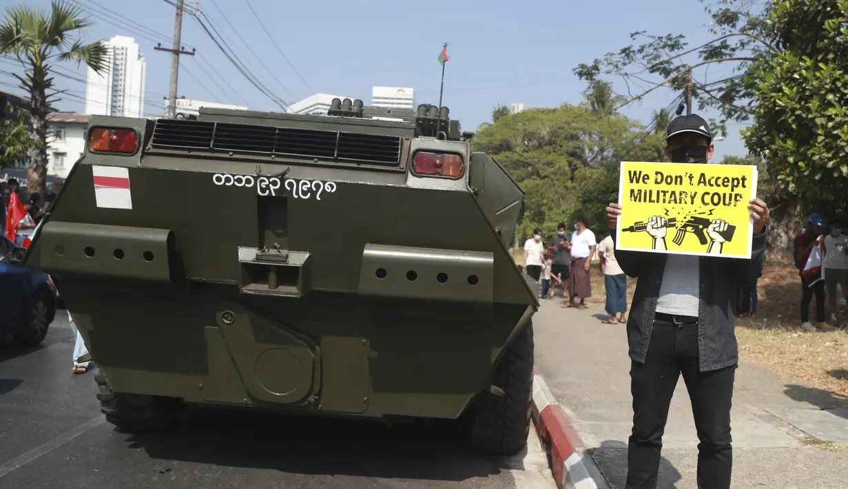 Seorang pengunjuk rasa anti-kudeta memegang poster di samping kendaraan lapis baja yang dikerahkan di luar gedung Bank Sentral Myanmar di Yangon, Senin (15/2/2021). Pasukan militer Myanmar mulai menerjunkan kendaraan lapis baja di kota-kota besar di Myanmar dan memutus akses internet. (AP Photo)