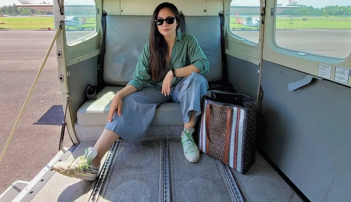 Berpose di dalam sebuah pesawat, Wulan Guritno tampil dengan nuansa liburan; kemeja hijau dan celana kain berwarna biru, yang dipadu dengan sneakers dan Burberry Monogram Stripe Print Tote Bag yang harganya diperkirakan sekitar Rp17,4 jutaan. Foto: Instagram.