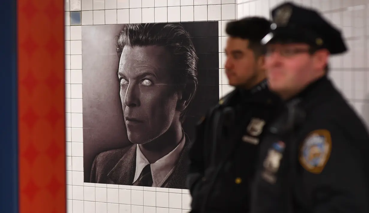 Dua petugas NYPD berdiri dekat instalasi seni yang memajang gambar David Bowie di stasiun kereta bawah tanah Broadway-Lafayette, New York City, 19 April 2018. Instalasi seni yang berlangsung hingga 13 Mei mendatang. (ANGELA WEISS/AFP)