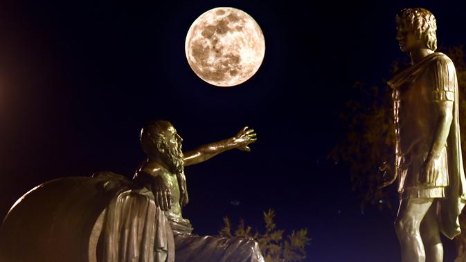 Fenomena super snow moon terlihat di antara patung Alexander Agung (kanan) dan Diogenes of Sinope (kiri) di Corinth, Yunani, Selasa (19/2). Super snow moon menampakkan diri di berbagai belahan dunia. (Valerie GACHE/AFP)