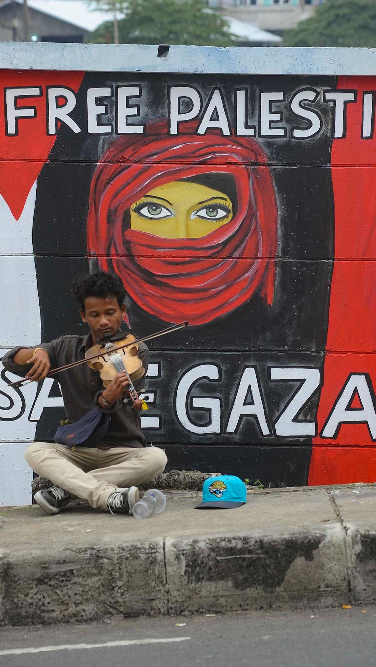 Seorang pengamen memainkan biola di depan mural "Free Palesine' di Jalan Juanda, Kota Depok, Jawa Barat, pada Rabu (8/11/2023).&nbsp;Merdeka.com/Arie Basuki