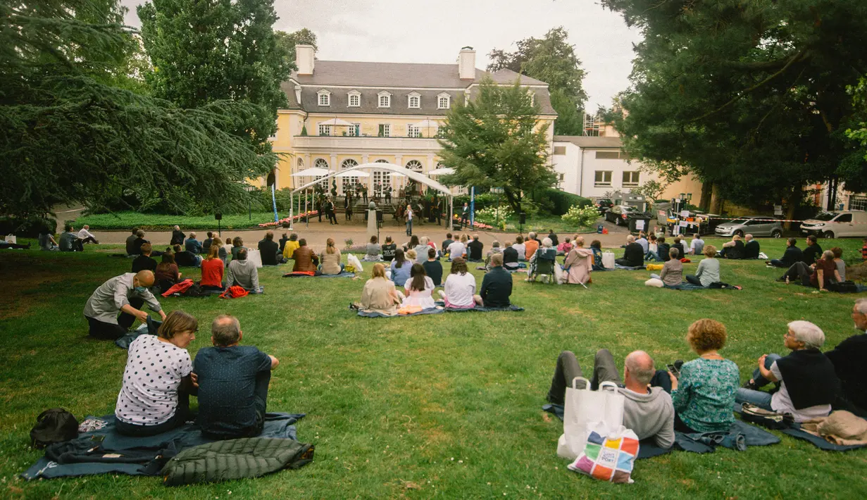 Warga menikmati konser yang diselenggarakan oleh Beethoven Orchestra Bonn di lapangan rumput sebuah taman di Bonn, Jerman, pada 28 Juni 2020. Penyelenggara menyediakan alas piknik bagi para audiens agar tetap menjaga jarak fisik di tengah kekhawatiran penyebaran COVID-19. (Xinhua/Tang Ying)