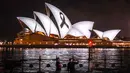 Orang-orang berdiri di depan pita hitam yang melambangkan kenangan dan duka cita, yang diproyeksikan ke Sydney Opera House, Australia, Kamis (9/2/2023). Gempa dahsyat berkekuatan 7,8 magnitudo terjadi di Turki dan Suriah pada Senin (6/2) lalu dan mengakibatkan kerusakan parah serta ribuan korban jiwa. (DAVID GRAY/AFP)