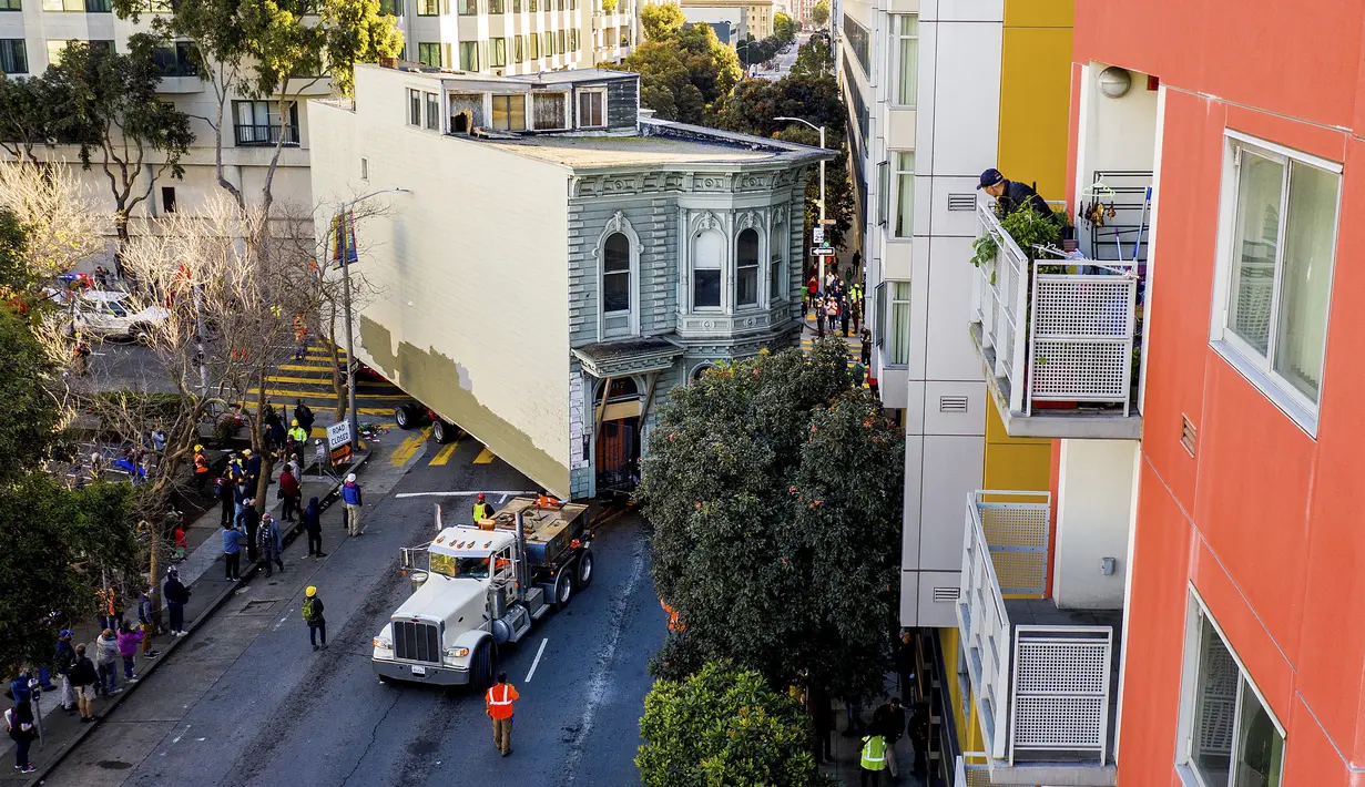 Seorang pria mengawasi dari balkon saat sebuah truk menarik rumah bergaya Victoria berusia 139 tahun di jalanan San Francisco, Minggu (21/2/2021). Rumah enam kamar tidut tersebut dipindahkan ke lokasi baru sekitar enam blok untuk memberi ruang bagi pengembangan kondominium. (AP Photo/Noah Berger)