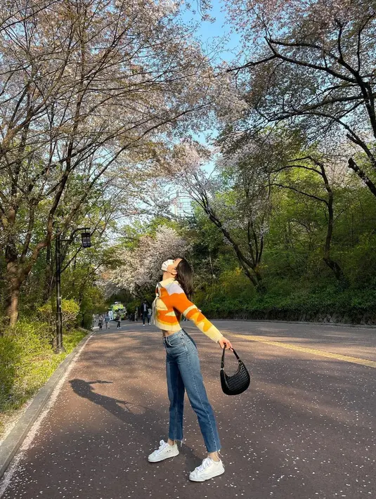 Berpose duduk di bawah pohon dengan bunga-bunga berguguran yang cantik, Lisa BLACKPINK terlihat mengenakan outfit bernuansa 80an, dengan sweater rajut berkerah, celana jeans, sneaker putih, dan mini bag hitam.