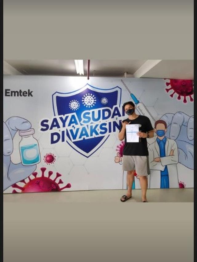 Kevin Kambey foto di depan backdrop “saya sudah divaksin” sambil memegang kartu vaksin.