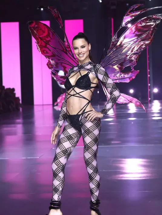 Ibu tiga anak, Adriana Lima tampil bersinar  memadukan bikini hitamnya dengan long pants dan cut out long sleeve top motif kotak-kotak. Ia melengkapi penampilannya dengan sayap hologram. [@runway.dolls].