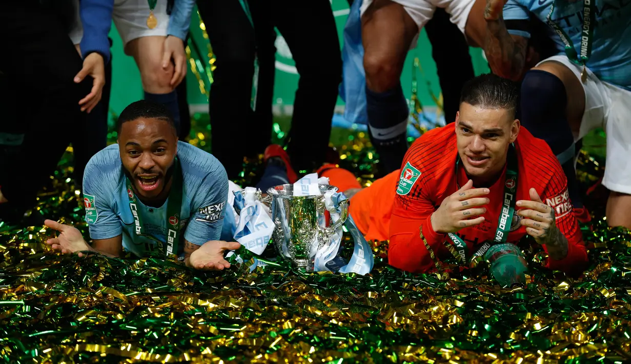 Gelandang dan kiper Manchester City, Raheem Sterling dan Ederson berbaring di dekat trofi setelah memenangkan final Piala Liga Inggris melawan Chelsea di Wembley, Minggu (24/2). Manchester City berhasil menjadi juara Piala Liga Inggris (Adrian DENNIS/AFP)