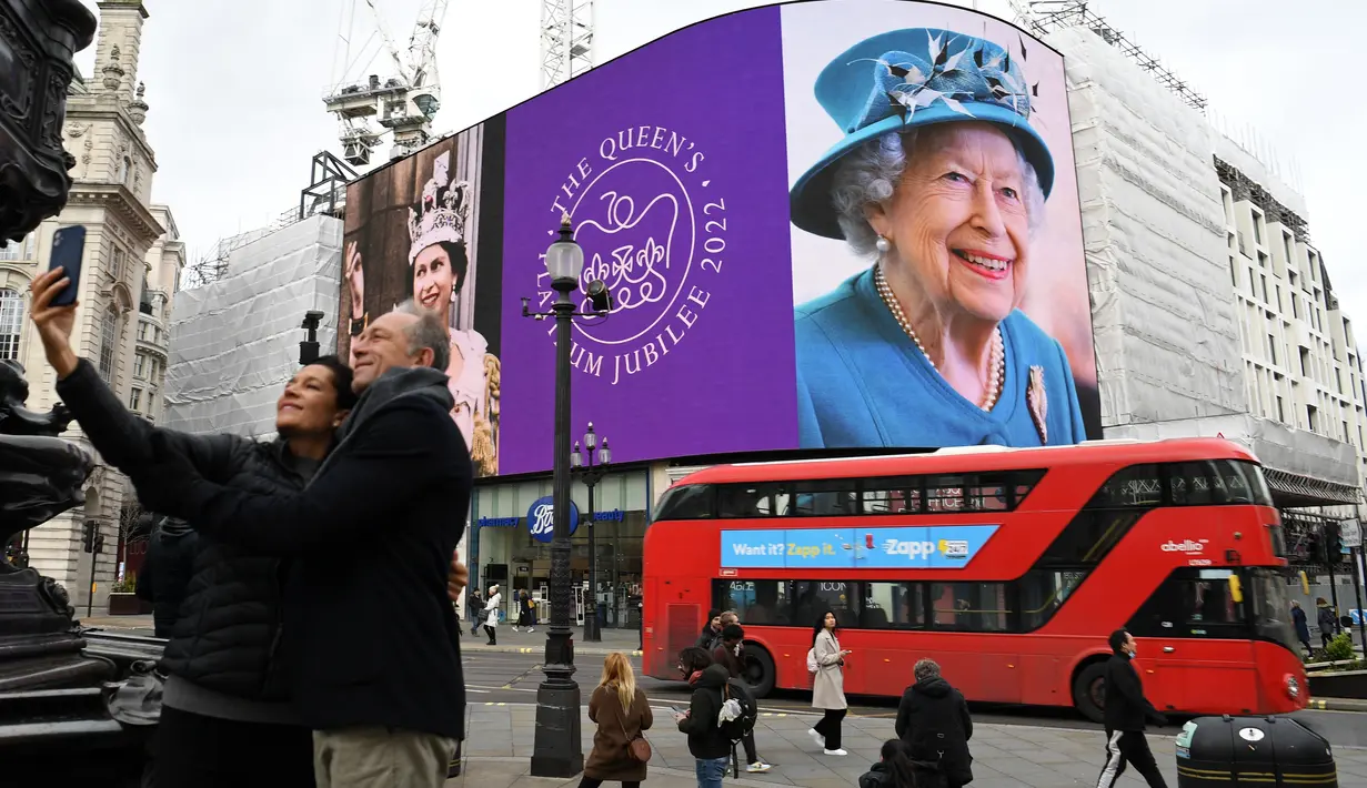 <p>Pasangan berfoto saat bus London merah lewat, di mana gambar Ratu Elizabeth II ditampilkan pada layar digital besar untuk menandai dimulainya Platinum Jubilee di Piccadilly Circus, 6 Februari 2022. Ratu Elizabeth II menjadi raja Inggris pertama yang memerintah selama tujuh dekade. (Daniel LEAL/AFP)</p>