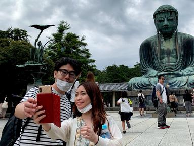 Sepasang pengunjung berswafoto dengan latar belakang The Great Buddha atau yang biasa disebut Kamakura Daibutsu di kuil Kotoku-in di Kamakura, Prefektur Kanagawa, Jepang, Sabtu (20/6/2020). Patung Buddha yang menjulang dengan tinggi 13,35 meter ini didirikan tahun 1252. (Behrouz MEHRI/AFP)