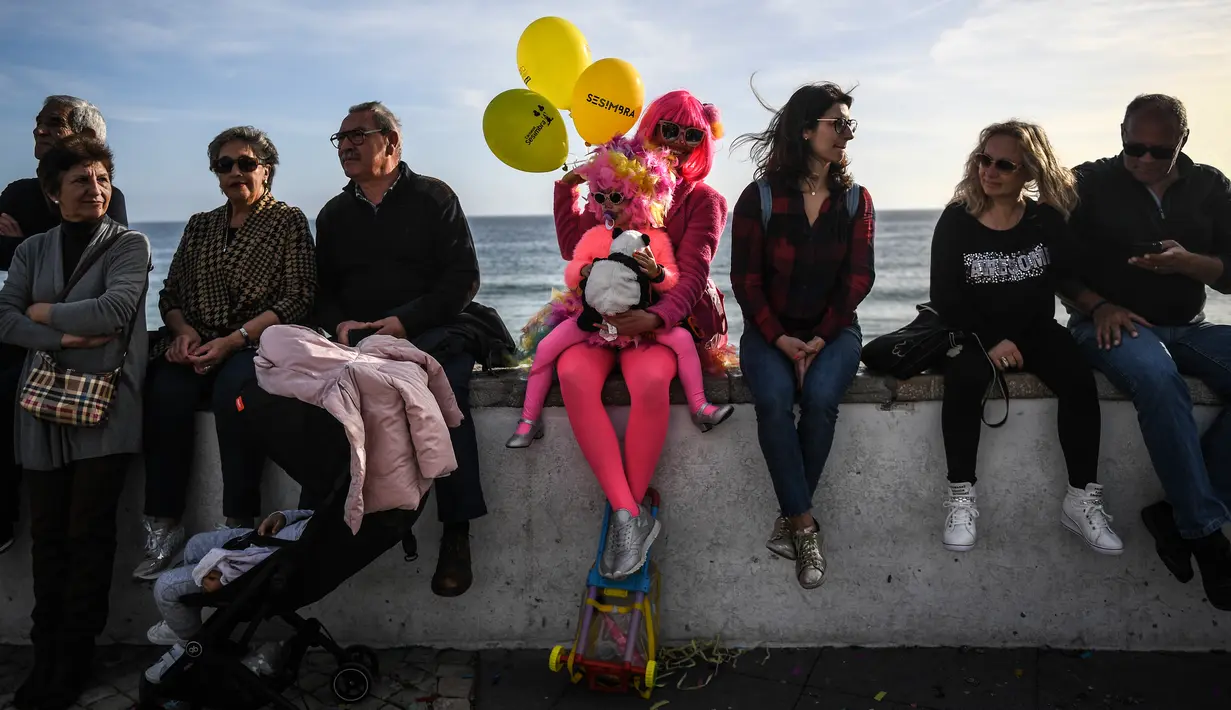 Seorang ibu dan anaknya dengan pakaian badut duduk bersama orang-orang yang menghadiri parade badut tahunan di Sesimbra, Portugal pada Senin (4/3/2019). Perayaan ini dilakukan setiap tahun dan jatuh pada hari Senin. (PATRICIA DE MELO MOREIRA / AFP)