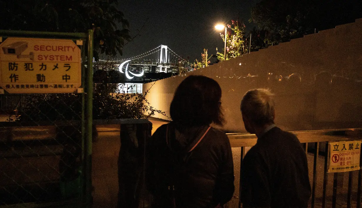 Warga melihat simbol Paralimpiade yang menyala dengan latar belakang Jembatan Pelangi di tepi Pantai Odaiba, Tokyo, Jepang, 23 Agustus 2021. Paralimpiade Tokyo 2020 mulai 24 Agustus 2021 sampai 5 September 2021. (Charly TRIBALLEAU/AFP)