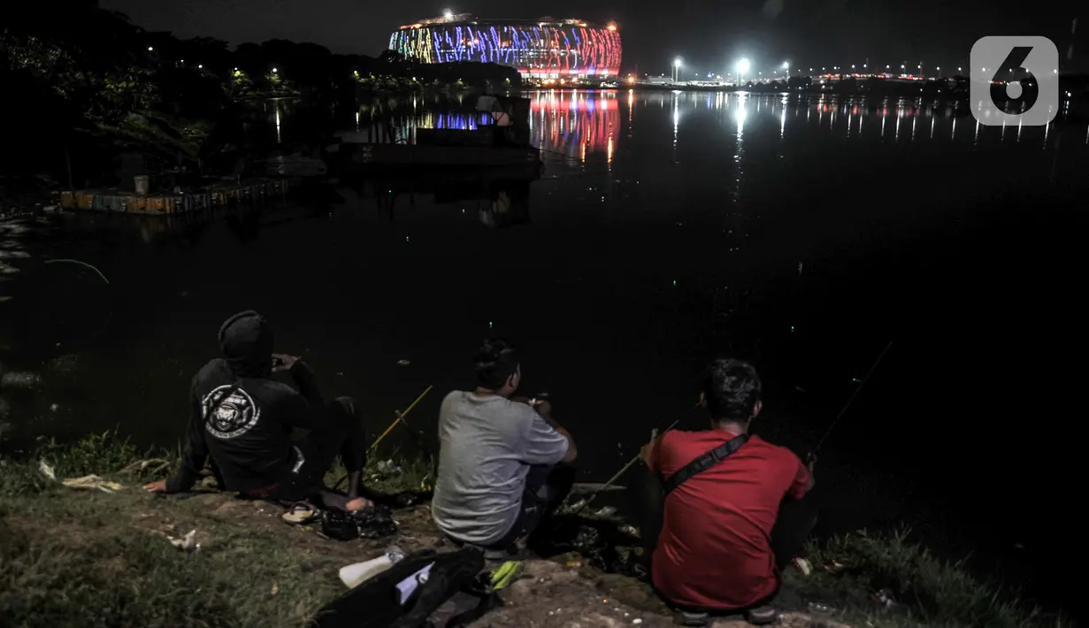 Warga memancing di Waduk Cincin dengan latar belakang Jakarta International Stadium (JIS), Papanggo, Jakarta Utara, Rabu (16/3/2022). Gemerlapnya warna-warni lampu stadion kebanggaan Ibu Kota ini menjadi daya tarik warga yang melintas di kawasan tersebut. (merdeka.com/Iqbal S. Nugroho)