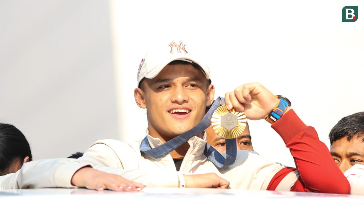 Peraih medali emas Olimpiade Paris 2024 dari cabang angkat besi, Rizki Juniansyah memamerkan medali saat melakukan pawai bersama sejumlah atlet dan ofisial tim Kontingen Indonesia lainnya menggunakan bus tingkat terbuka Transjakarta menuju Istana Negara, Jakarta Pusat, Kamis (15/8/2024). (Bola.com/Abdul Aziz)