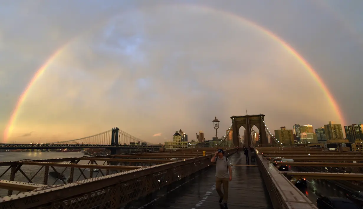 Pejalan kaki melintasi Jembatan Brooklyn saat pelangi menghiasi langit New York City, AS (15/5). Jembatan Brooklyn adalah salah satu jembatan suspensi tertua di Amerika Serikat. (AFP Photo/Hector Retamal)