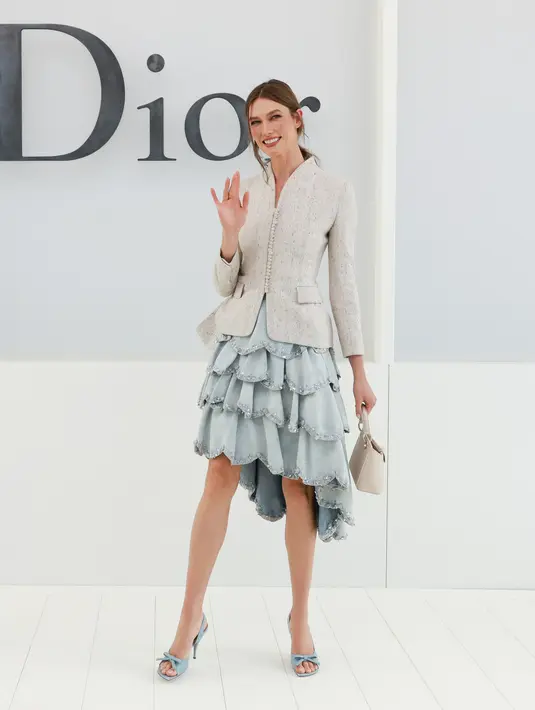 Karlie Kloss tampil ceria dengan blazer krem berkilau dipadukan rok tiered berlapis warna biru dusty yang playful. Ia tak ragu melambaikan tangan dengan tas mini krem [dok. Dior]