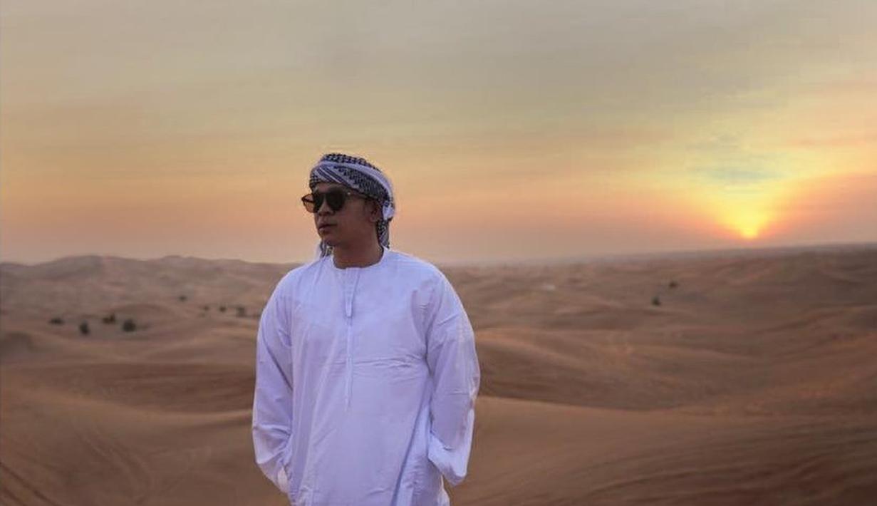 Selain pernyataannya yang bikin heboh, gaya berpakaian Jordi Onsu juga menjadi sorotan. Ia sempat tampil mengenakan jubah gamis putih lengkap dengan sorban bermotif hitam-putih, menciptakan kesan elegan khas Timur Tengah. Dengan latar belakang padang pasir dan matahari terbenam, penampilannya semakin dramatis dan menambah spekulasi di kalangan netizen.  (Liputan6.com/IG/@jordionsu)