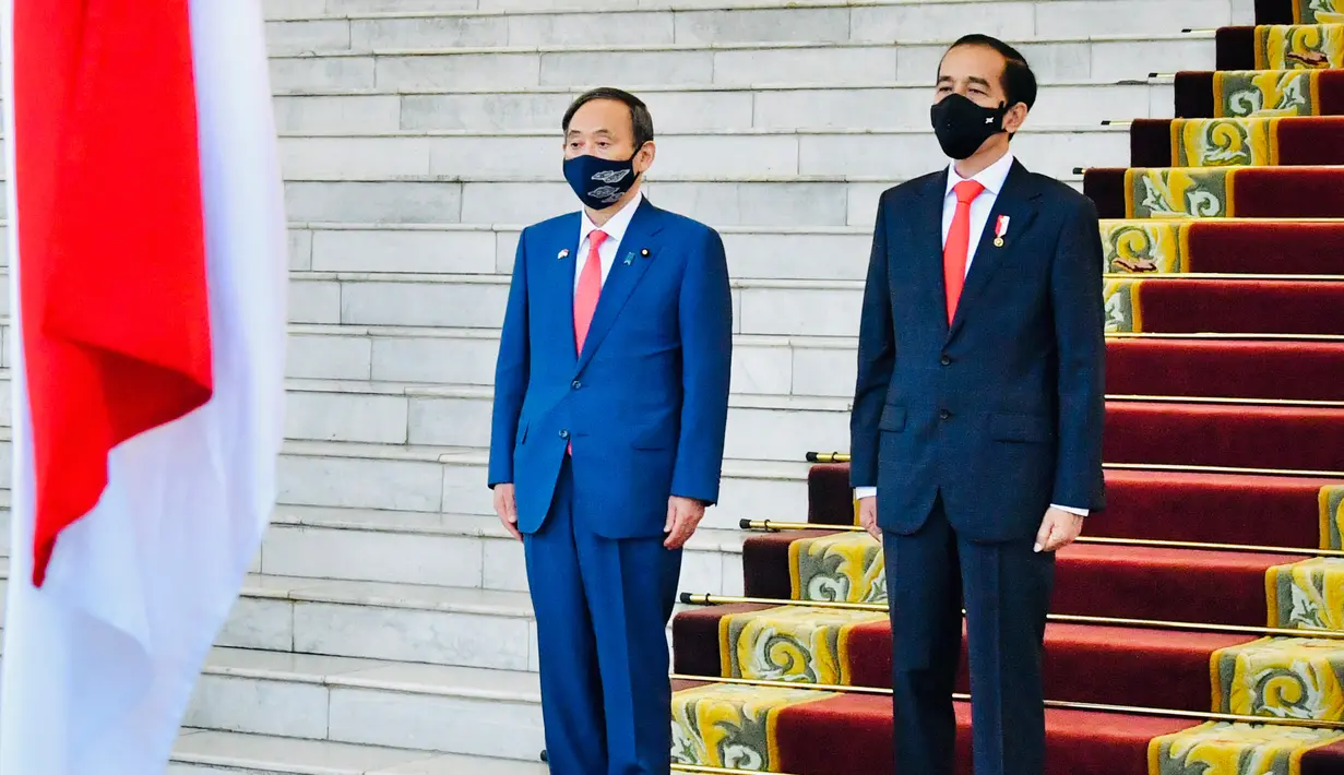 Presiden Joko Widodo bersama Perdana Menteri Jepang Yoshihide Suga (kiri), sama-sama mengenakan masker sebagai upaya mencegah penyebaran Covid-19, mendengarkan lagu kebangsaan saat upacara penyambutan di Istana Bogor, 20 Oktober 2020. (Laily Rachev/Indonesian Presidential Palace via AP)
