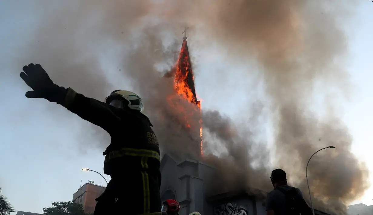 Petugas pemadam kebakaran berdiri di depan gereja La Asuncion yang terbakar saat puluhan ribu orang berunjuk rasa di Kota Santiago, Chile, Minggu (18/10/2020). Demonstrasi itu digelar untuk memperingati satu tahun protes besar menuntut kesetaraan di Chile. (AP Photo/Esteban Felix)