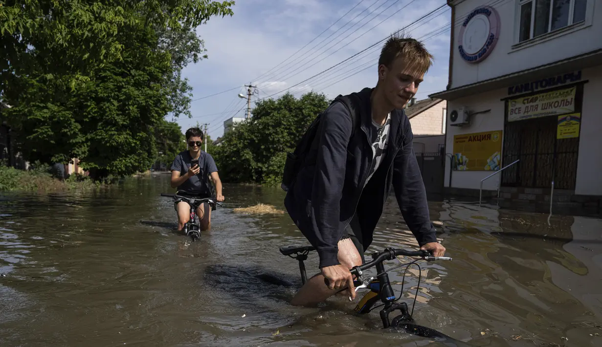<p>Penduduk setempat mencoba mengendarai sepeda mereka di sepanjang jalan yang banjir setelah bendungan Kakhovka meledak semalaman, di Kherson, Ukraina, Selasa, 6 Juni 2023.(AP Photo/Evgeniy Maloletka)</p>