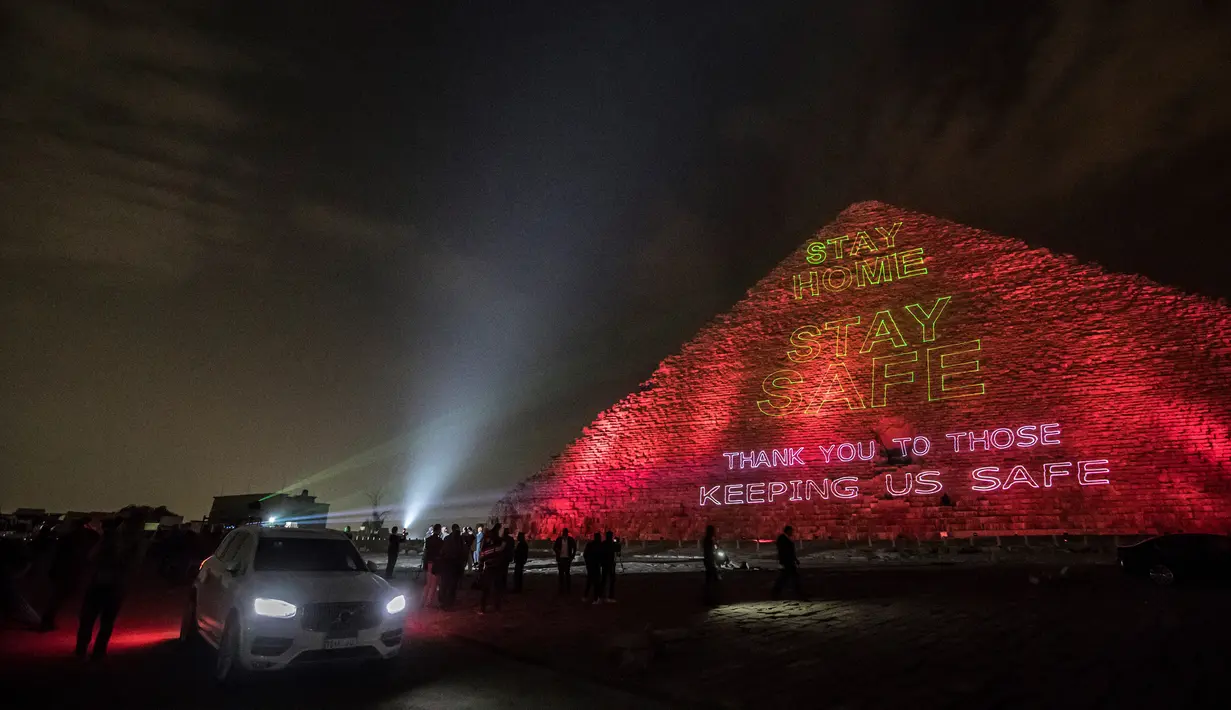 Orang-orang mengambil gambar piramida besar Kheops yang memproyeksikan tulisan "Stay home, thanks to those keeping us safe" di Giza, Mesir, Senin (30/3/2020). Piramida tersebut menampilkan pesan persatuan dan solidaritas bagi mereka yang memerangi virus corona di seluruh dunia. (Khaled DESOUKI/AFP)