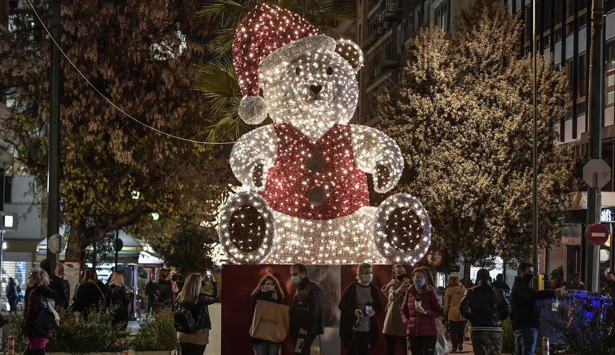 Orang-orang berdiri di dekat beruang raksasa yang diterangi dengan dekorasi Natal di jalan komersial utama Athena, Yunani pada 21 Desember 2020. Yunani akan merayakan musim Natal di bawah penguncian wilayah atau lockdown akibat virus corona hingga 7 Januari 2021 mendatang. (LOUISA GOULIAMAKI/AFP)