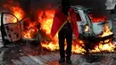 Seorang demonstran berselimut bendera Albania berdiri di depan mobil yang terbakar saat aksi unjuk rasa antipemerintah di luar kantor Perdana Menteri di Tirana, Albania, Sabtu (17/10/2026). (AP Photo/Hameraldi Agolli)