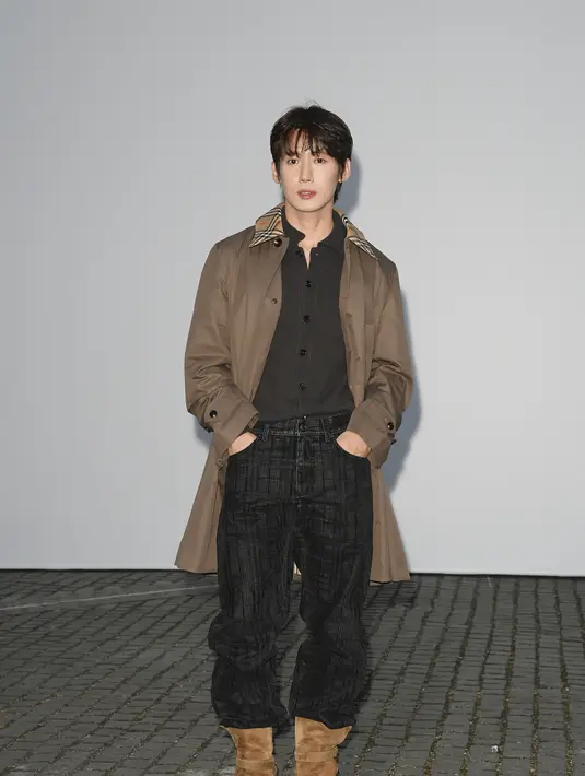 Seung Ju Han mengenakan trench coat cokelat dengan kerah bermotif kotak-kotak khas Burberry, dipadukan sweater rajut gelap, celana plaid gelap, dan boots suede cokelat. [dok. Burberry]