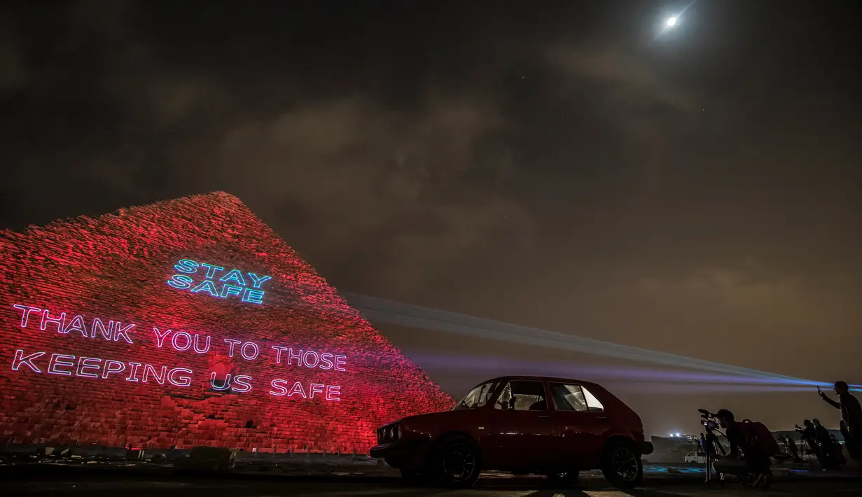 Orang-orang mengambil gambar piramida besar Kheops yang memproyeksikan tulisan "Stay home, thanks to those keeping us safe" di Giza, Mesir, Senin (30/3/2020). Piramida tersebut menampilkan pesan persatuan dan solidaritas bagi mereka yang memerangi virus corona di seluruh dunia. (Khaled DESOUKI/AFP)