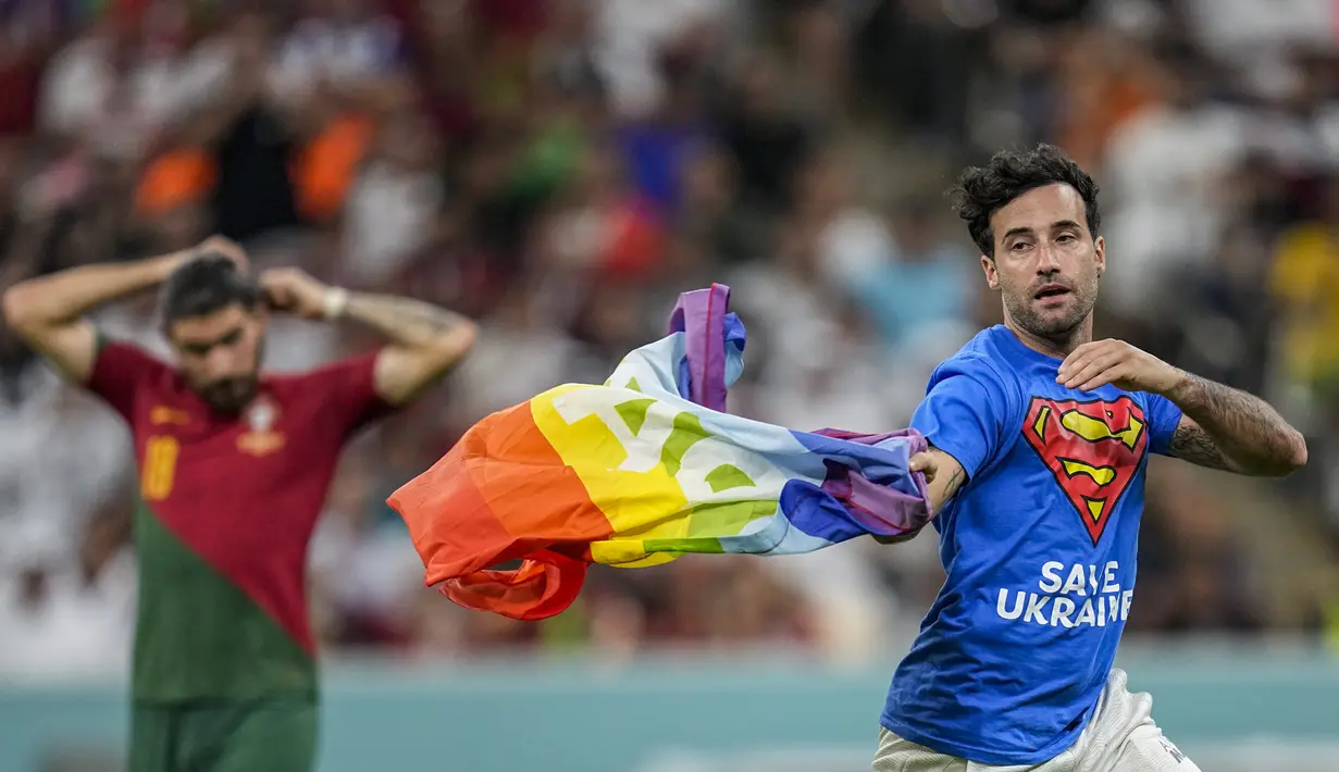 Seorang pria membawa bendera pelangi yang identik dengan LGBT berlari melintasi lapangan selama pertandingan grup H Piala Dunia 2022 Qatar antara Portugal melawan Uruguay di Stadion Lusail di Lusail, Qatar, Senin, 28 November 2022.  Pria yang diketahui bernama Mario Ferri tersebut juga mengenakan kaos yang bertuliskan protes terhadap perang Ukraina dan Rusia yang telah berlangsung kurang lebih Sembilan bulan, serta di bagian belakang kaos bertuliskan “Hormati Wanita Iran”. (AP Photo/Abbie Parr)