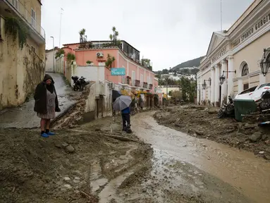 Orang-orang berjalan melalui lumpur di jalan yang banjir setelah hujan deras memicu tanah longsor yang meruntuhkan bangunan dan menyebabkan sebanyak 12 orang hilang, di Casamicciola, di pulau selatan Italia Ischia, Sabtu, 26 November 2022. Petugas pemadam kebakaran sedang melakukan upaya penyelamatan karena bala bantuan sedang dikirim dari Napoli terdekat, tetapi mengalami kesulitan dalam mencapai pulau baik dengan perahu motor atau helikopter karena cuaca. (AP Photo/Salvatore Laporta)