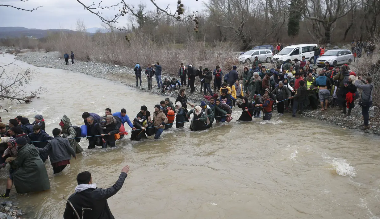 Ribuan migran menyeberangi sungai dengan berpegangan pada seutas tali di perbatasan Yunani-Makedonia, Idomeni, Yunani (14/3/2016). Lebih dari 1.000 migran nekat menembus sungai untuk mencari jalan keluar perbatasan. (Reuters/Stoyan Nenov)