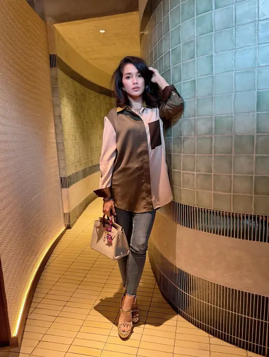 Gaya cantik Ussy pakai kemeja satin dua warna; cokelat dan pink, dipadu celana jeans abu-abu, ia juga mengenakan alas kaki tinggi dan menenteng tas kecokelatan yang senada. [Foto: Instagram/ussypratama]