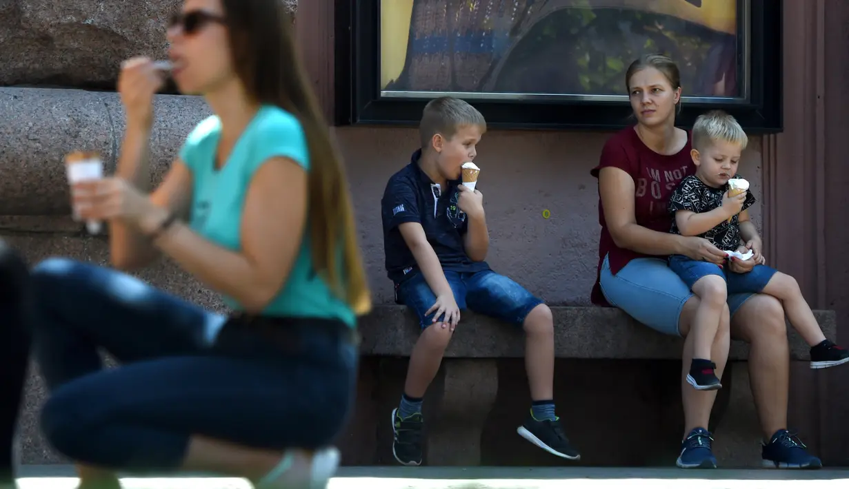 Dua anak didampingi ibunya menikmati es krim selama gelombang panas di ibukota Ukraina (21/8/2019). Panasnya cuaca yang ekstrem membuat anak-anak menyejukkan diri mereka di bawah guyuran air mancur. (AFP Photo/Sergei Supinsky)