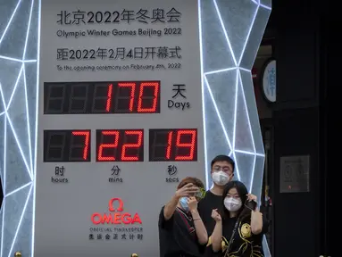 Orang-orang yang memakai masker untuk melindungi diri dari COVID-19 berpose di depan layar yang menunjukkan jam hitung mundur ke Olimpiade Musim Dingin 2022 di Beijing (18/8/2021).  Pejabat tinggi Beijing menegaskan kembali perlunya anti-pencegahan yang ketat. (AP Photo/Mark Schiefelbein)