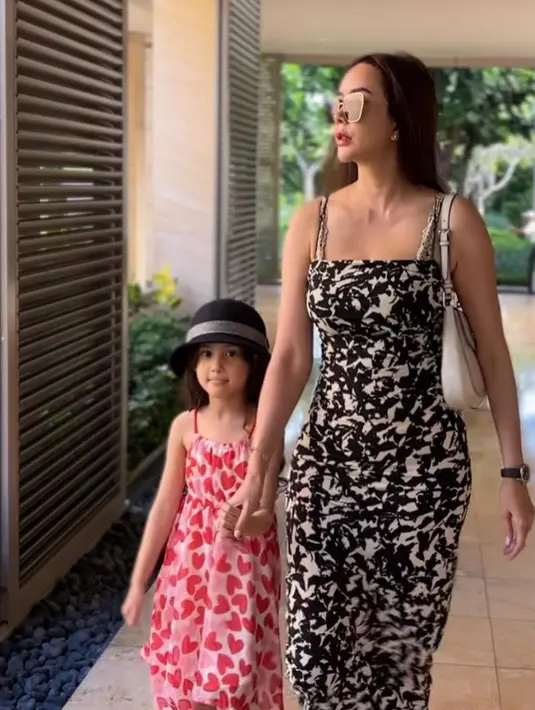 Aura Kasih yang sedang berjalan bersama putrinya, Arabella. Aura Kasih terlihat anggun mengenakan gaun panjang bermotif hitam dan putih, serta kacamata hitam yang menambah kesan elegan. (Liputan6.com/IG/@aurakasih)