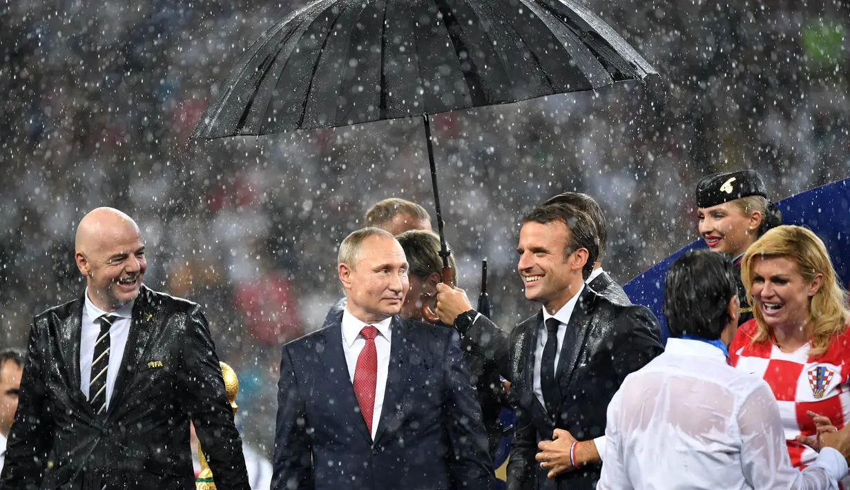 Presiden Rusia Vladimir Putin dilindungi oleh seorang pemandu yang memegang payung sementara para pejabat lainnya basah kuyup saat hujan turun selama penyerahan medali dan trofi Piala Dunia 2018 di Stadion Luzhniki, Minggu (15/7). (Kirill KUDRYAVTSEV/AFP)