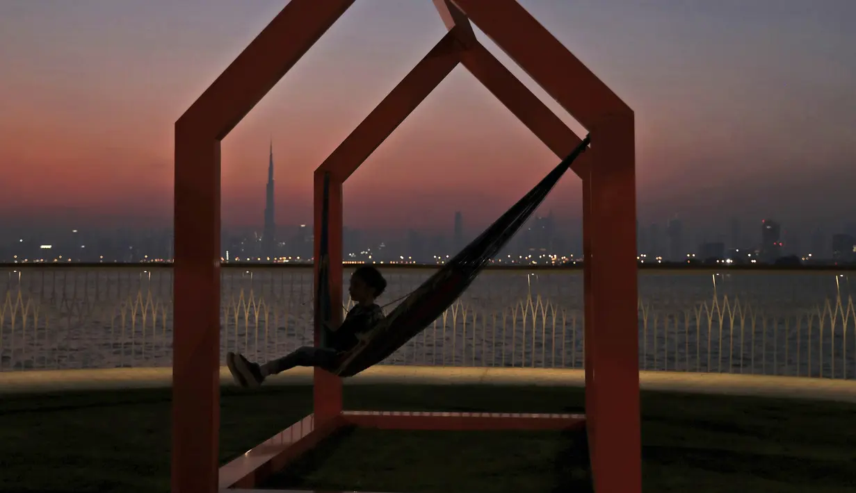 Seorang gadis berayun ketika matahari terbenam di belakang cakrawala kota dengan latar gedung tertinggi di dunia Burj Khalifa, di Dubai, Uni Emirat Arab (18/10/2019). (AP Photo/Kamran Jebreili)