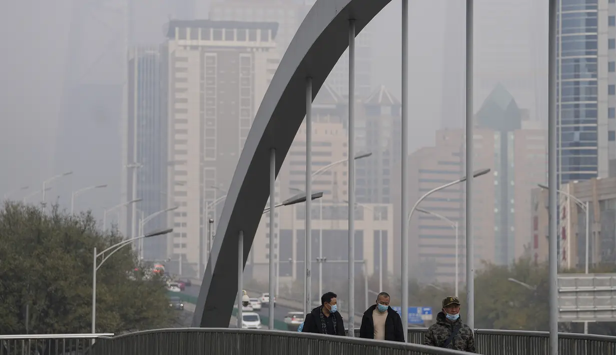 Orang-orang yang memakai masker untuk membantu mengekang penyebaran virus corona berjalan di atas jembatan di atas gedung perkantoran yang diselimuti kabut polusi di Beijing, Kamis (18/11/2021).  (AP Photo/Andy Wong)