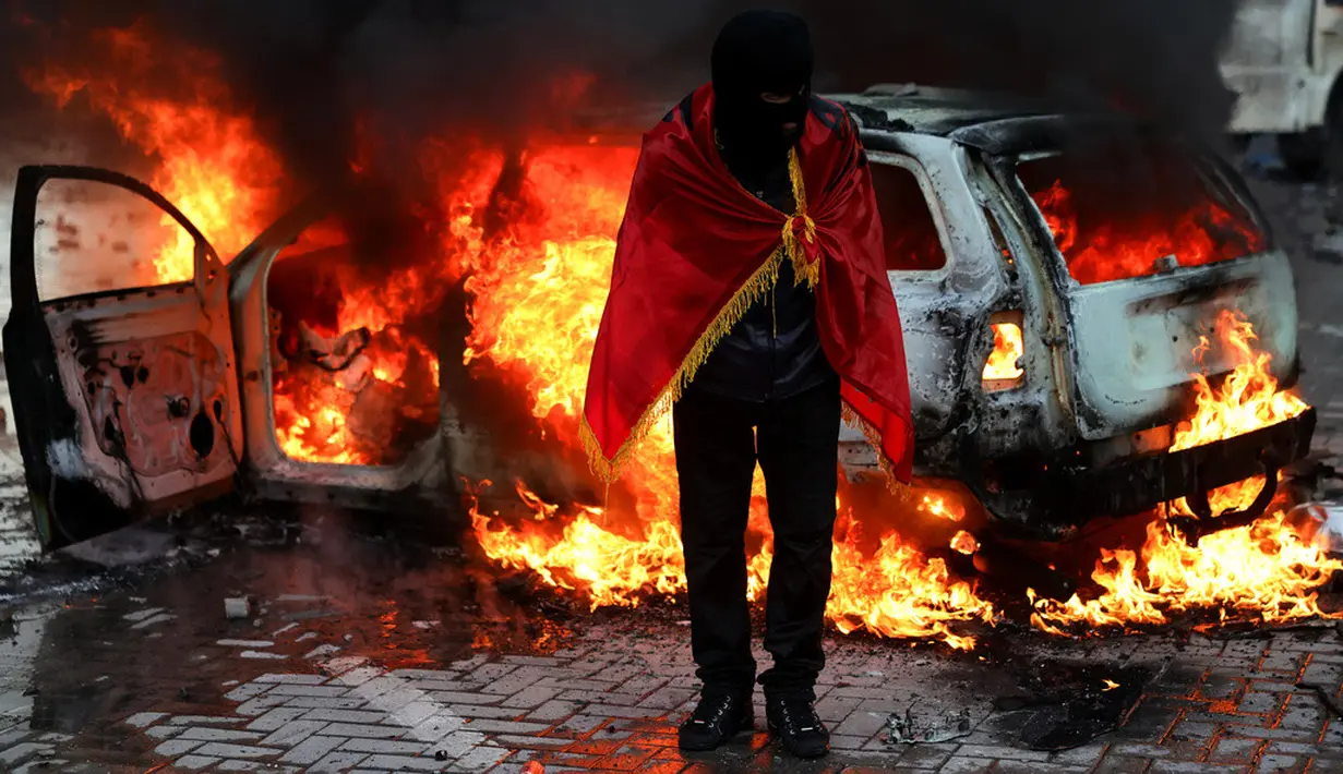Seorang demonstran berselimut bendera Albania berdiri di depan mobil yang terbakar saat aksi unjuk rasa antipemerintah di luar kantor Perdana Menteri di Tirana, Albania, Sabtu (17/10/2026). (AP Photo/Hameraldi Agolli)