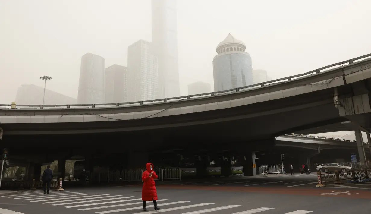 Seorang wanita menyeberang jalan dekat gedung pencakar langit saat badai pasir di Beijing (28/3/2021). Badai pasir kembali menyapu Beijing pada Minggu (28/3), mengubah langit menjadi kuning, membatasi jarak pandang dan mengganggu lalu lintas. (AP Photo/Ng Han Guan)