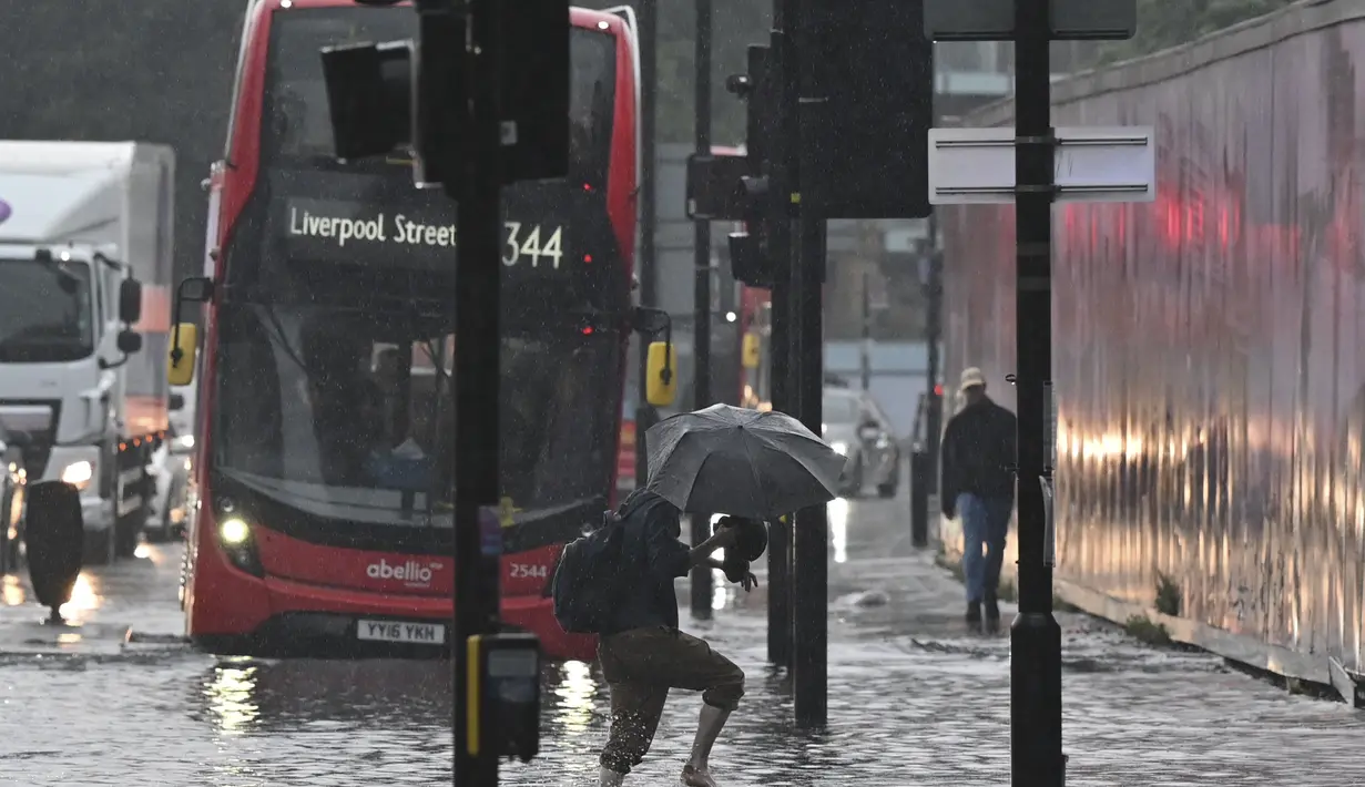 Seorang pejalan kaki melintasi perairan dalam di jalan yang banjir di distrik The Nine Elms London(25/7/2021). Bus dan mobil terjebak banjir ketika jalan-jalan di London banjir pada hari Minggu, ketika badai petir berulang kali melanda ibu kota Inggris. (AFP/Justin Tallis)