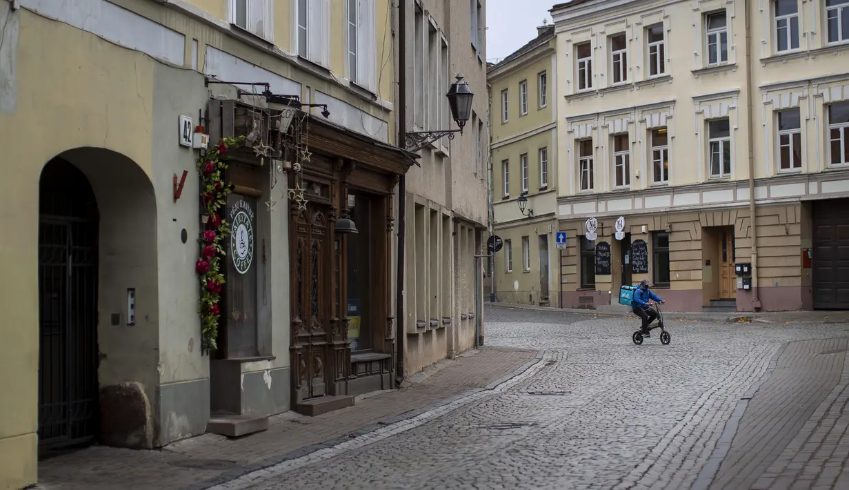 Seorang kurir pengiriman makanan yang mengenakan masker untuk mencegah penyebaran virus corona, mengendarai sepeda di Kota Tua, Vilnius, Lithuania (7/11/2020). Penguncian dilakukan untuk membendung penyebaran virus corona. (AP Photo/Mindaugas Kulbis)