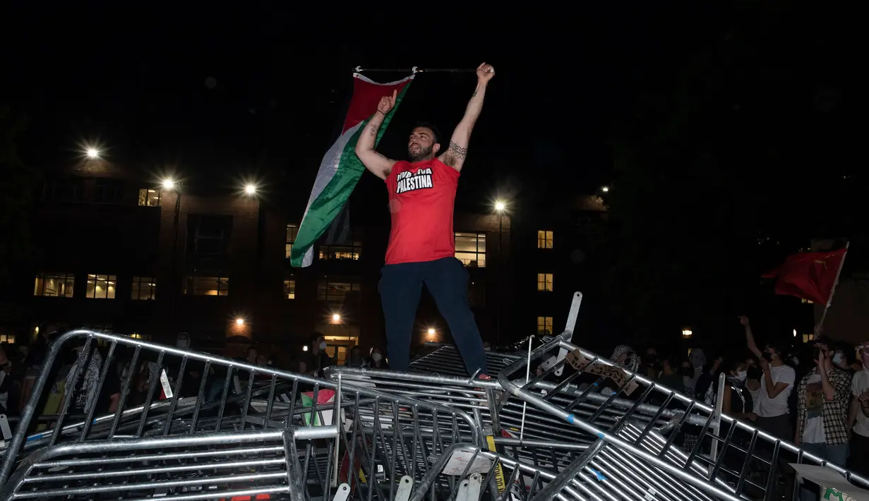 Seorang mahasiswa Universitas George Washington melambaikan bendera Palestina saat berdiri di atas barikade polisi yang diterobos untuk menduduki University Yard dalam aksi protes pro-Palestina di Washington, Minggu, 28 April 2024. (AP Photo/Cliff Owen)